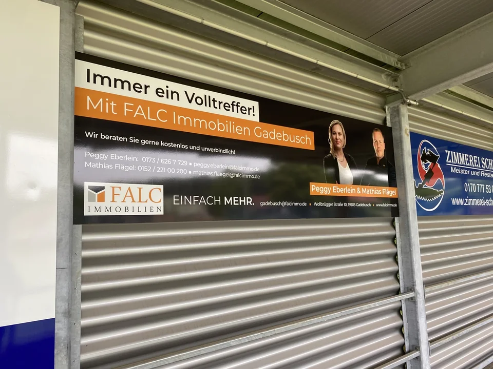 Schwarz-orange Werbetafel für FALC Immobilien Gadebusch an einer Metallwand