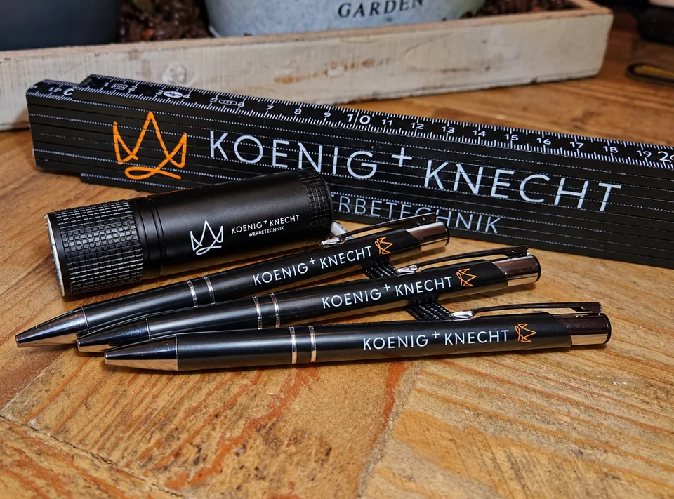Schwarze Kugelschreiber, Taschenlampe und Zollstock mit KOENIG+KNECHT-Logo auf Holz